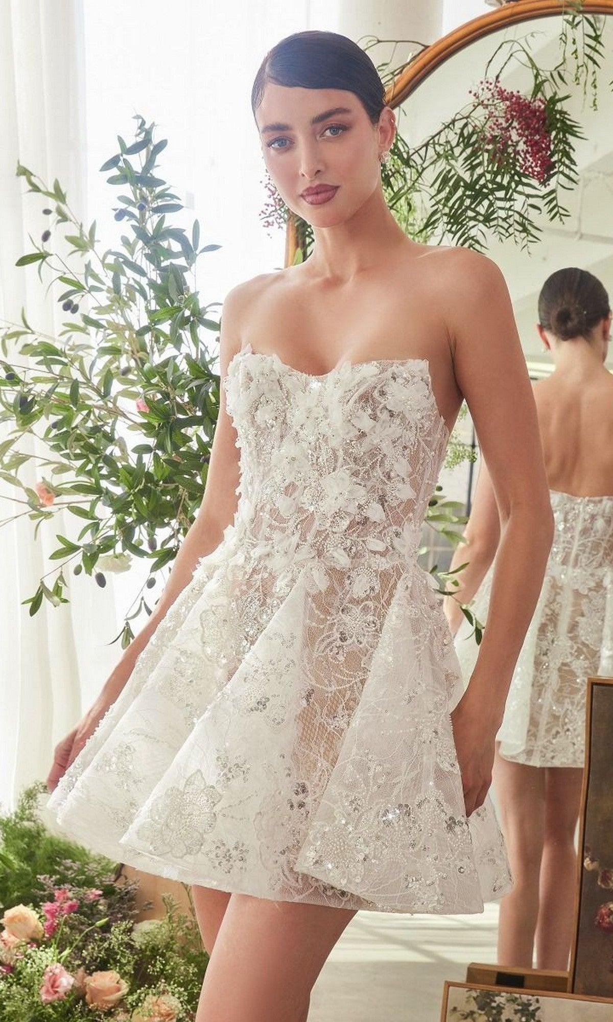A woman stands indoors wearing the Andrea and Leo WL082 short white dress, a strapless fit and flare style with lace floral appliques; greenery and a mirror add elegance to the scene.